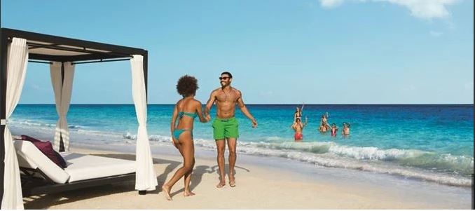Couple walking together along a sandy beach beside the ocean while several people enjoy the water nearby and a beach cabana sits along the shore.