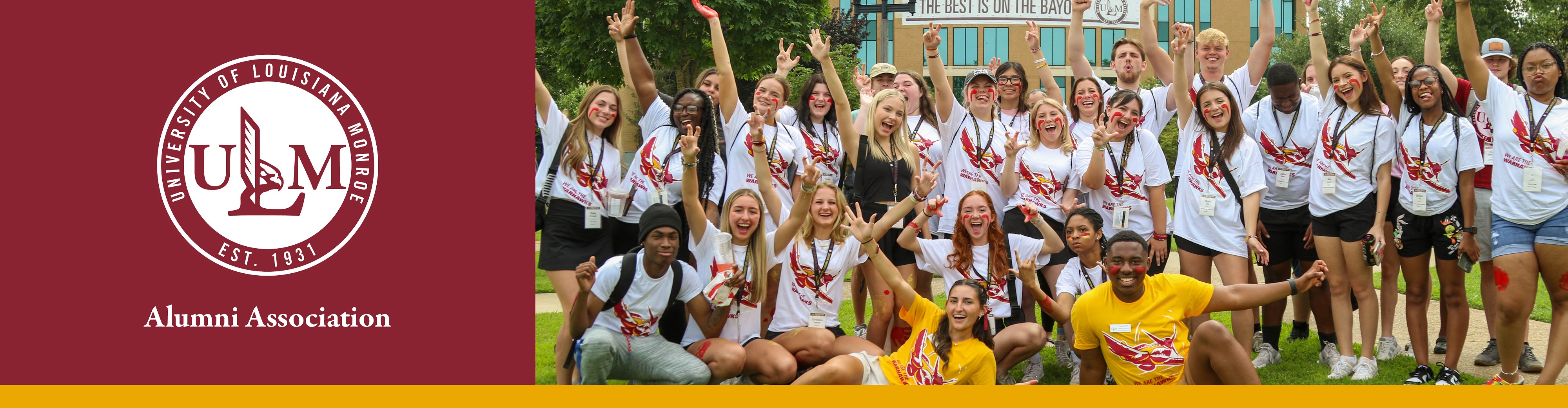 A group of college students cheer and smile at the camera. They are wearing matching shirts and stand in front of the ULM library.