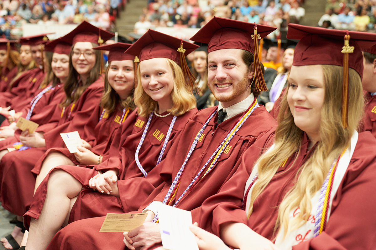 Row of students graduating seated, all wearing cap and gowns, audience in the background