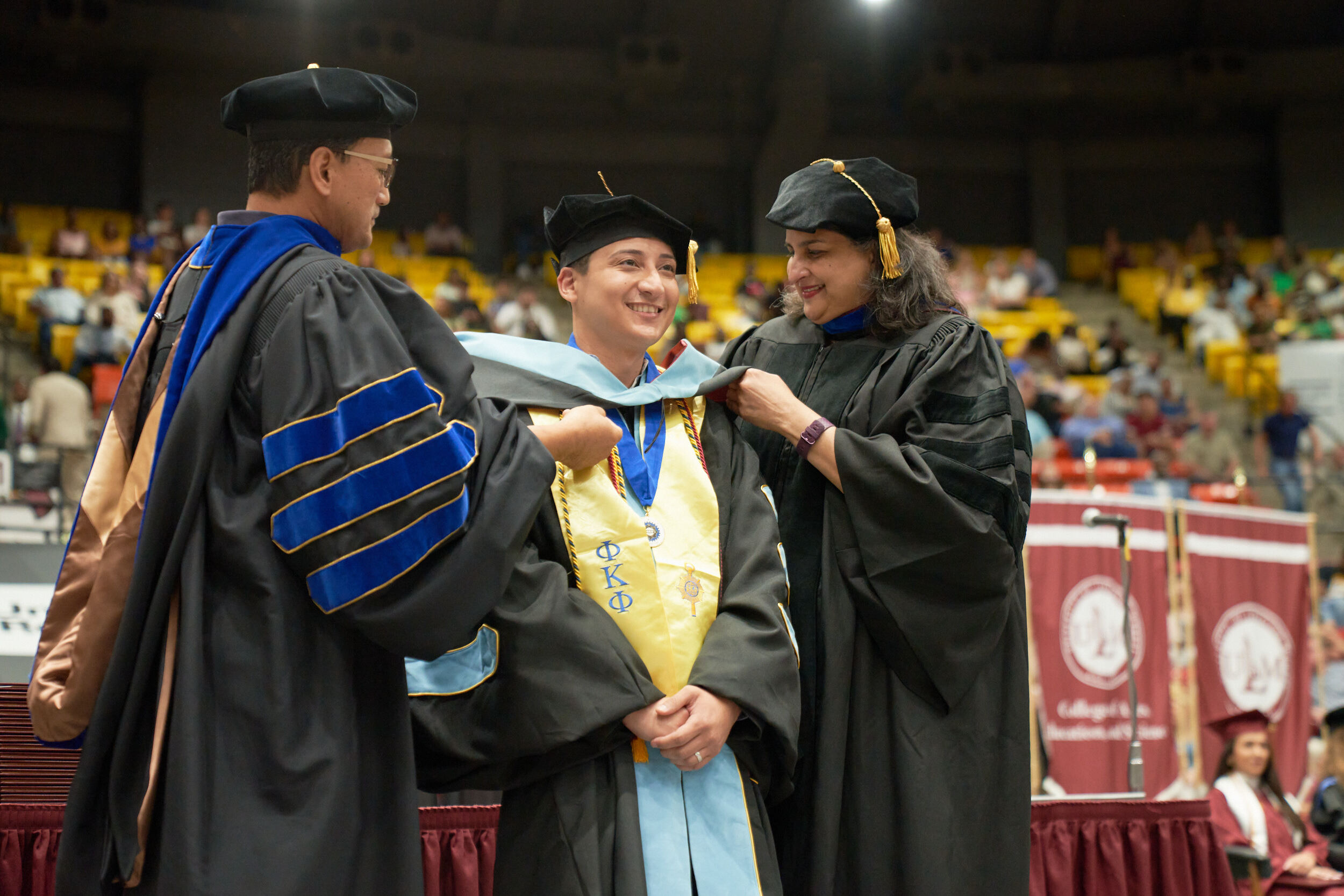 Doctoral hood being placed over head of male student by dean and professor, all are smiling, audience in background