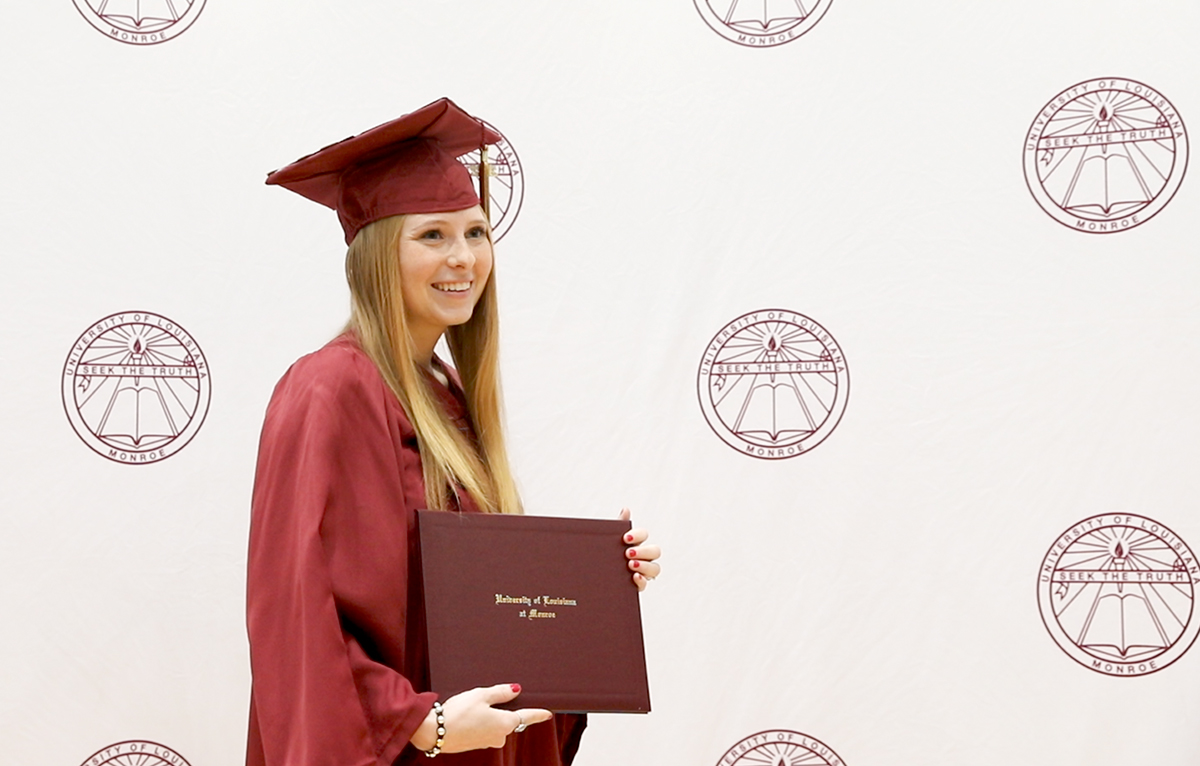 Student smiling and posing with degree in front of backdrop at ceremony 