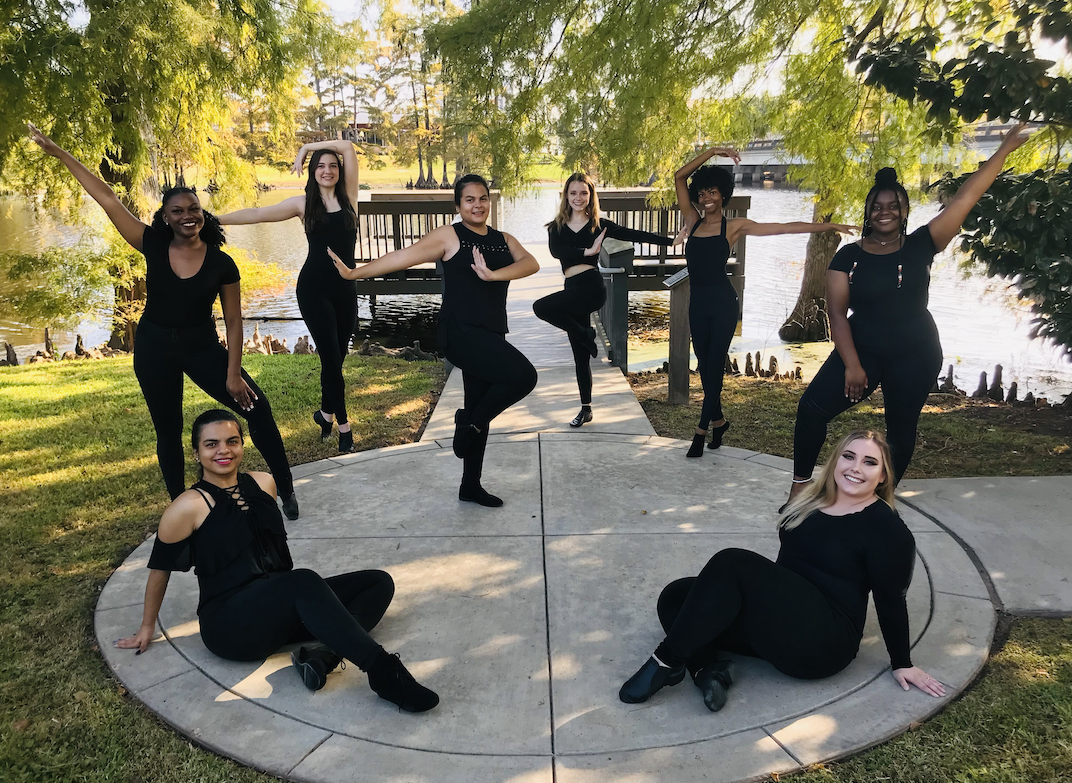Members of the ULM Repertory Dance Ensemble are wearing black performance clothes and posing outside in front of Bayou DeSiard.