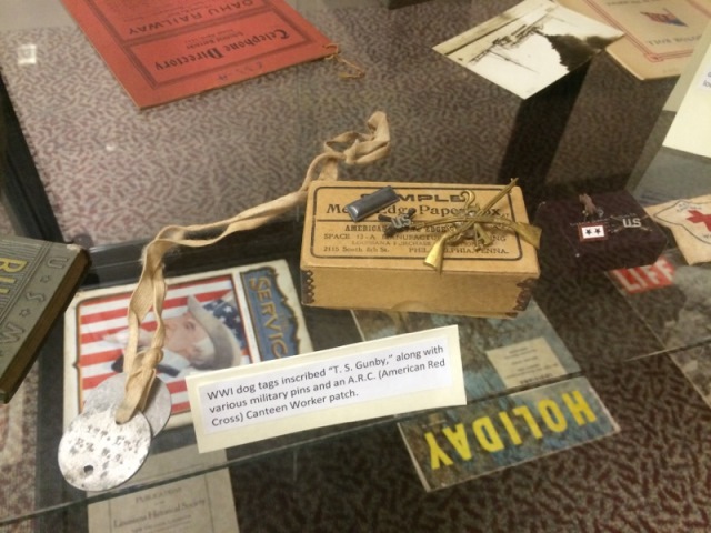 The centerpiece is a collection of WWI dog tags inscribed with "T. S. Gunby," accompanied by various military pins and an American Red Cross (A.R.C.) Canteen Worker patch.