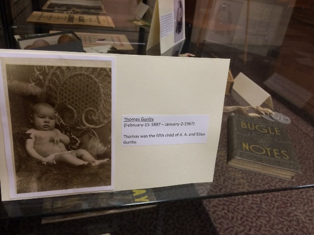 The displayed photograph features an infant, identified as Thomas Gunby. Thomas lived from February 15, 1897, to January 2, 1967. He was the fifth child of A.A. and Ellen Gunby. A book titled "BUGLE NOTES" is also visible in the display.