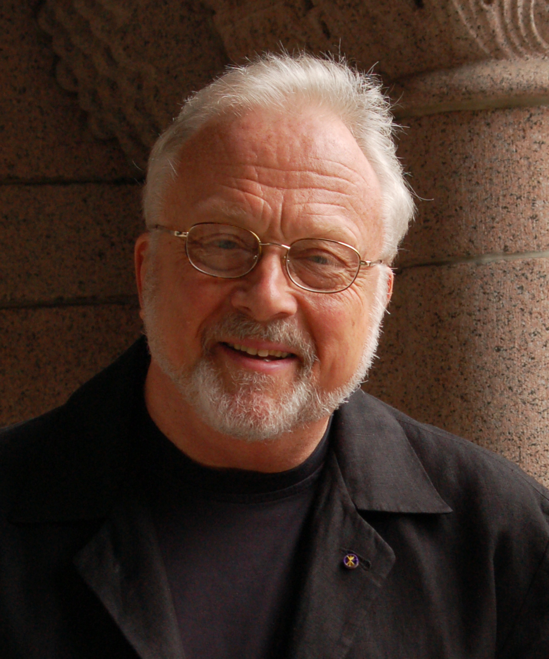 Composer William Bolcolm is standing in front of a stone building and wearing black clothes.