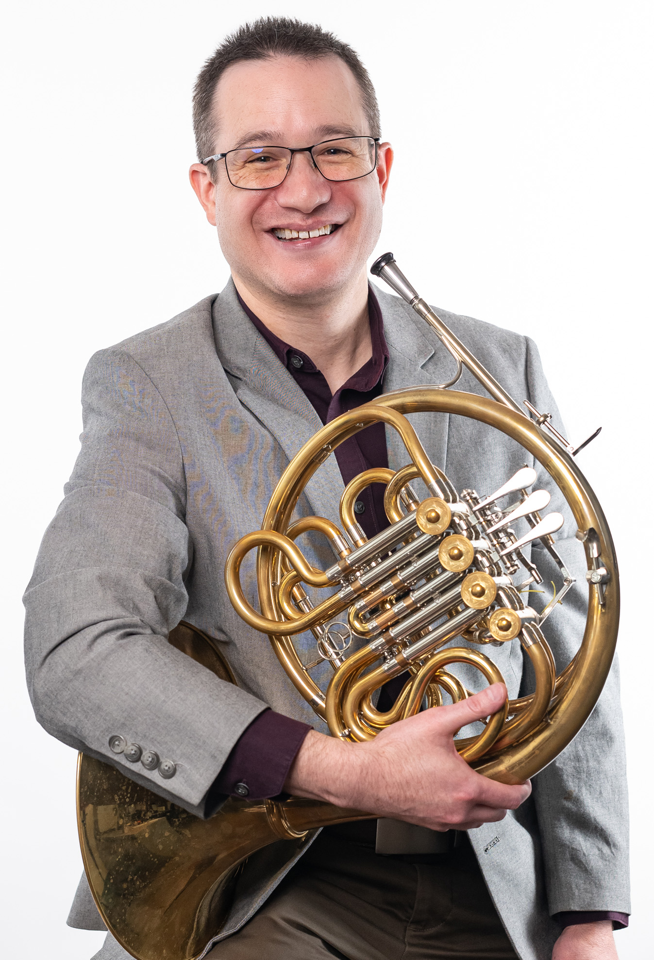 James Boldin, Professor of Music, is holding a French horn and wearing a grey jacket and a maroon shirt.
