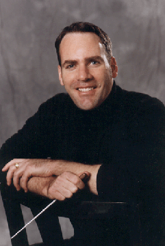 Composer David Childs is seated and holding a baton.