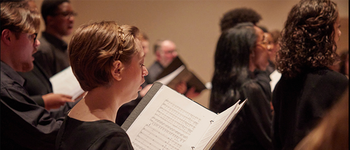 Members of a ULM choral ensemble perform on stage in Biedenharn Recital Hall. A singer is holding a black folder of music and the pages are visible. 