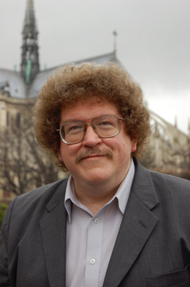 Composer Eric Ewazen is wearing a gray jacked and standing in front of Notre Dame Cathedral in Paris.