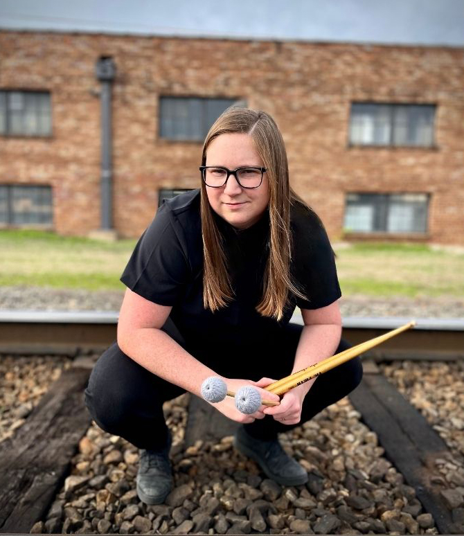 Dr. Kathryn Irwin is holding drumsticks and mallets, and is posing on a railroad track in front of a red brick building. She is wearing glasses and black clothes.