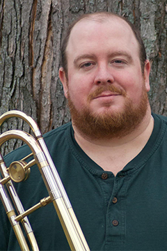 Adam Johnson is holding a trombone and standing in front of a tree. He has a beard and is wearing a green shirt.