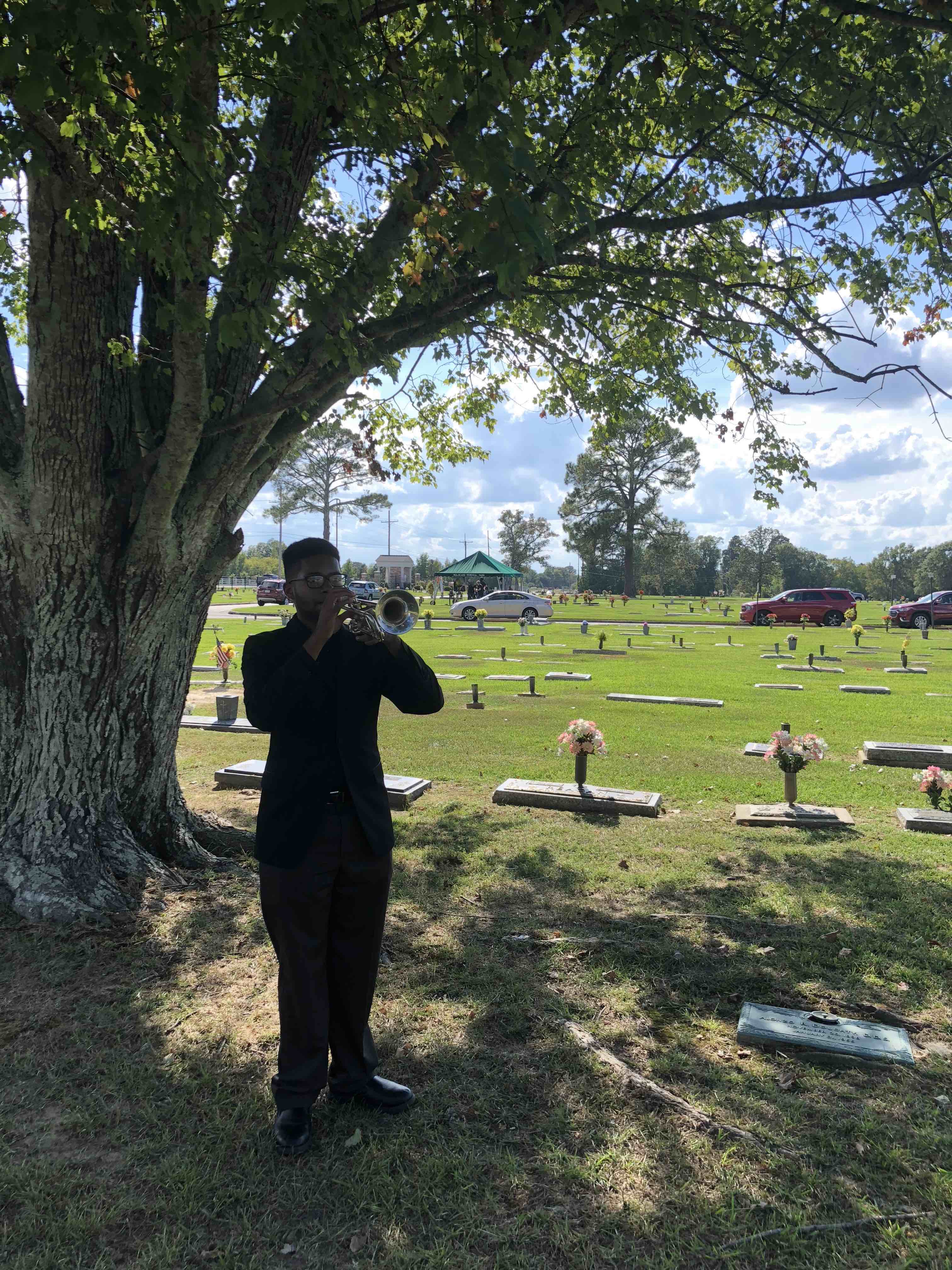 A trumpet player is standing outside underneath a tree in a cemetery. The trumpet player is wearing black clothes. 