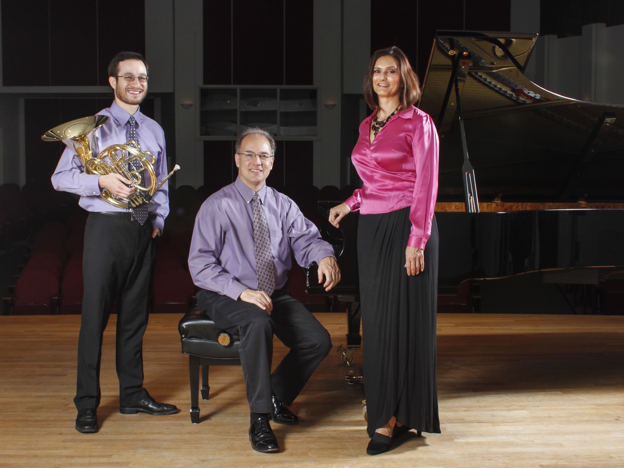 James Boldin is holding a French horn, Richard Seiler is seated at a grand piano with the lid open, and Claire Vangelisti is standing beside the piano. Boldin and Seiler are wearing purple shirts, and Vangelisti is wearing a pink shirt. 