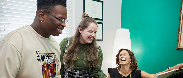 Dr. Vangelisti is seated at a piano, and two students are standing beside her. All three are smiling.