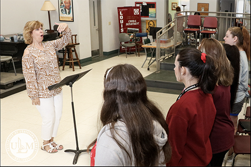 A conductor standing in front of a choir of women in a Biedenharn Hall classroom.