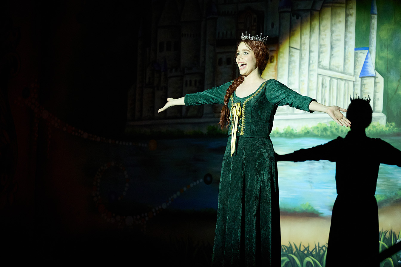 In this scene from Shrek the Musical, the student portraying Princess Fiona is wearing a green dress and standing alone on stage and singing with her arms spread out.